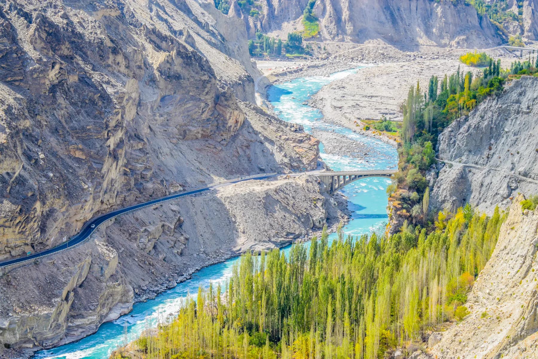 The turquoise Hunza River between steep rock walls, with a road and bridge crossing the valley and tall trees along the bank.