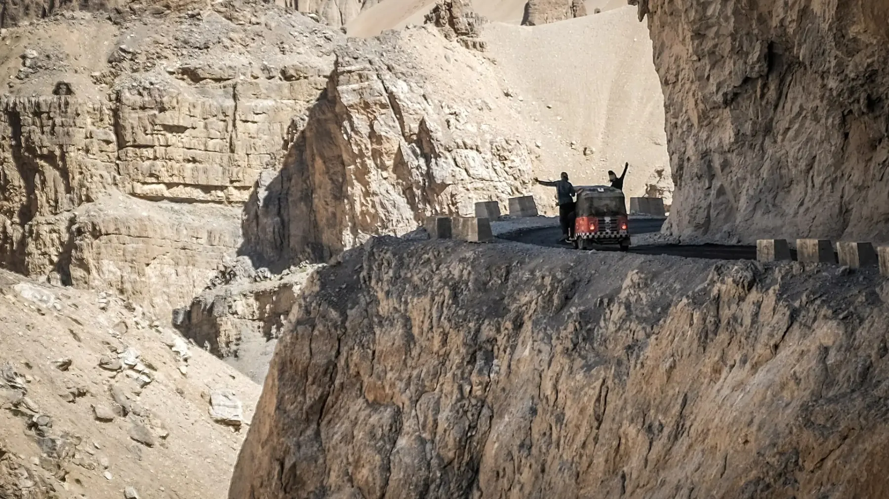 A tuk-tuk stopped on a narrow cliff road cut into an enormous face of pale eroded rock, two figures beside it with their arms raised.
