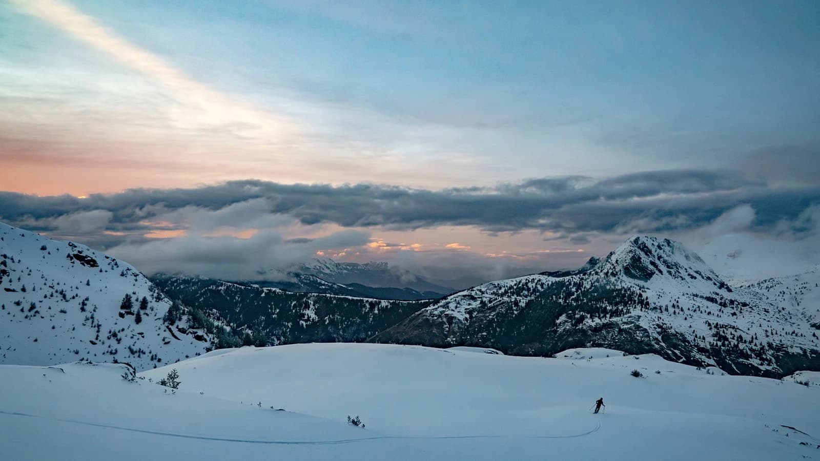 Dusk over a wide snow plateau in the Accursed Mountains, a sea of cloud filling the valleys below and a single skier small against the slope.
