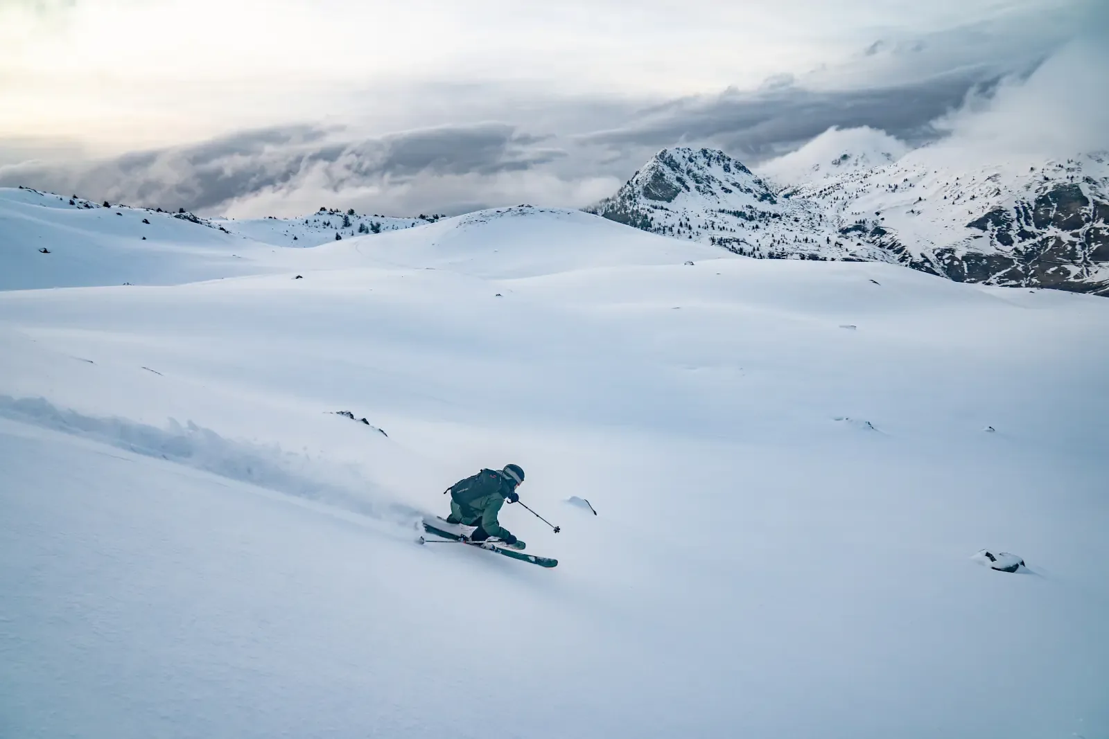 A skier turns across an open snowy slope below a rocky summit, spraying snow from the skis.