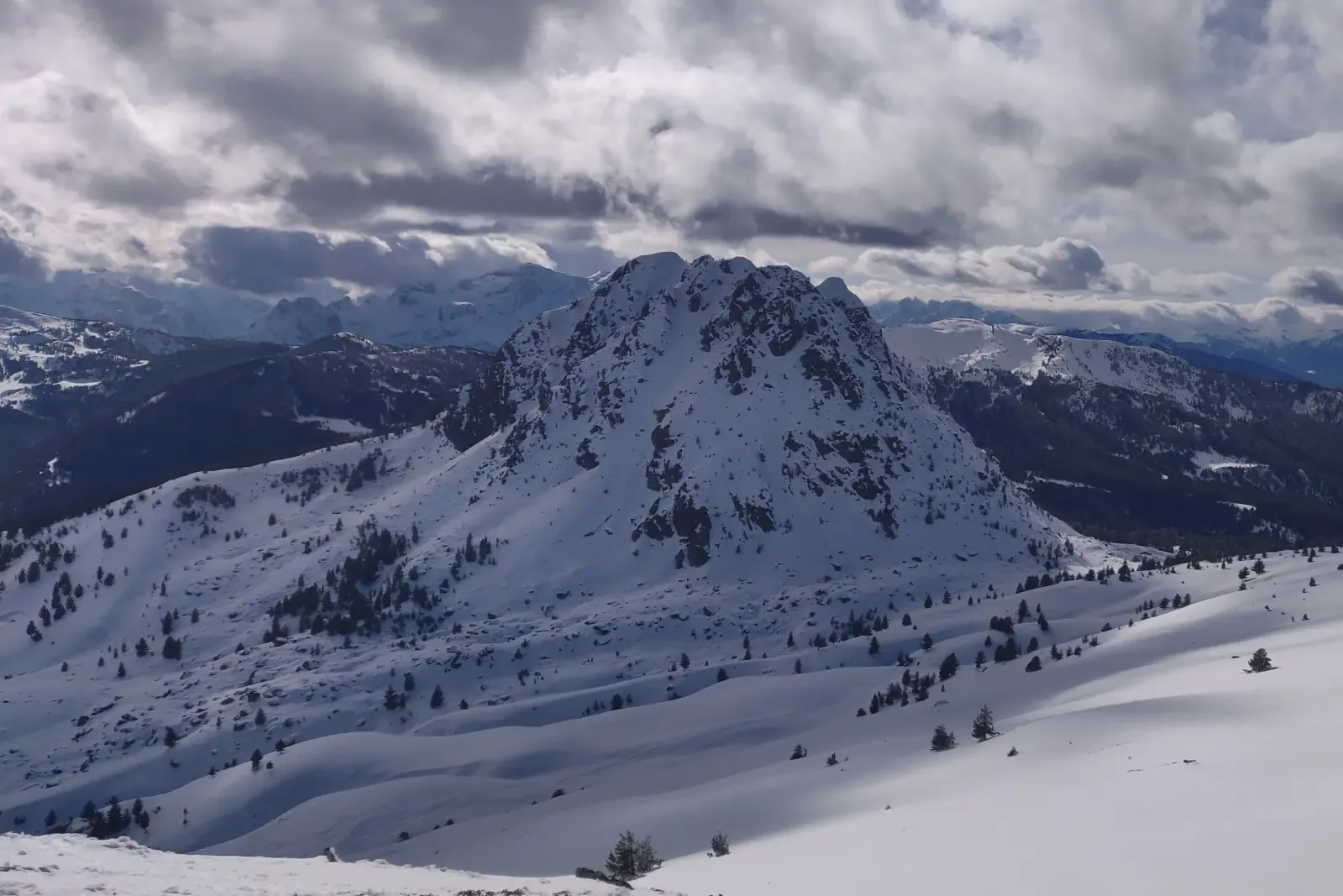 A rocky peak rises above an open snow basin scattered with trees, with further mountain ridges under broken cloud.