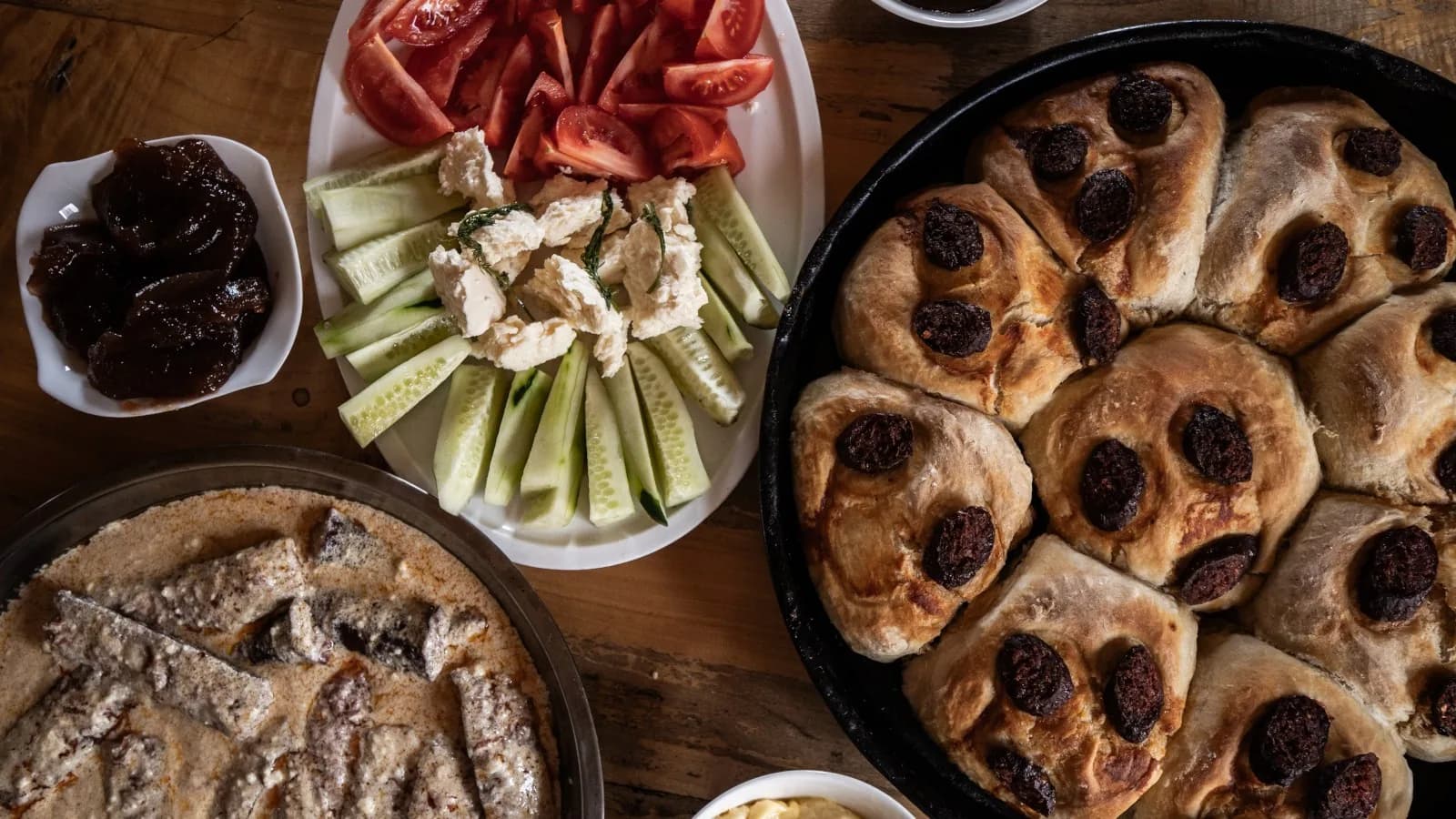 Looking down on a wooden table: a black pan of baked bread rolls each topped with a dark round slice, a metal dish of a pale sliced pie, a plate of cut tomatoes, cucumber batons and crumbled white cheese, and a bowl of dark preserves.