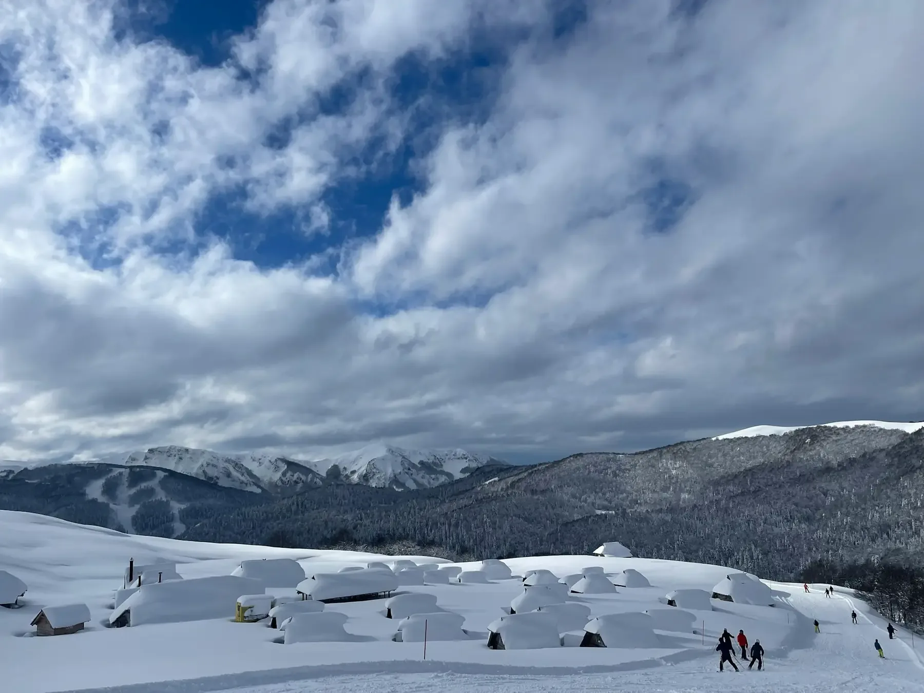 Low wooden huts sit deep in snow beside a track with people on it, below wooded ridges and heavy clouds.