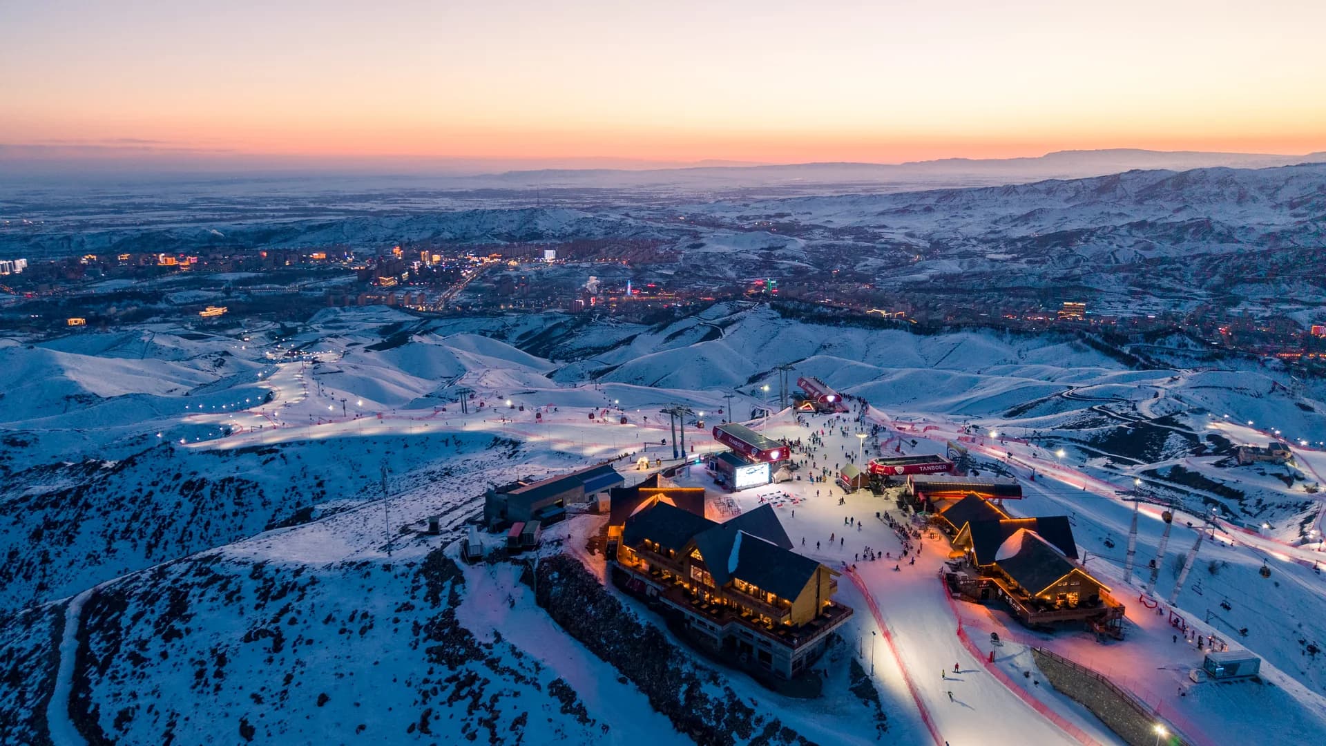 Snow-covered ski slopes and buildings are seen above a city at blue hour.