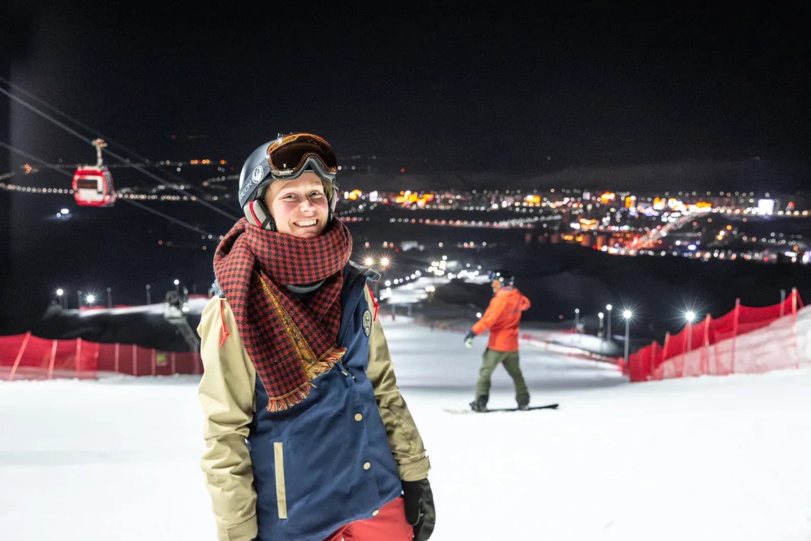 A smiling woman in a helmet with goggles raised, a red scarf and a blue-and-cream jacket stands on a floodlit slope with city lights below.