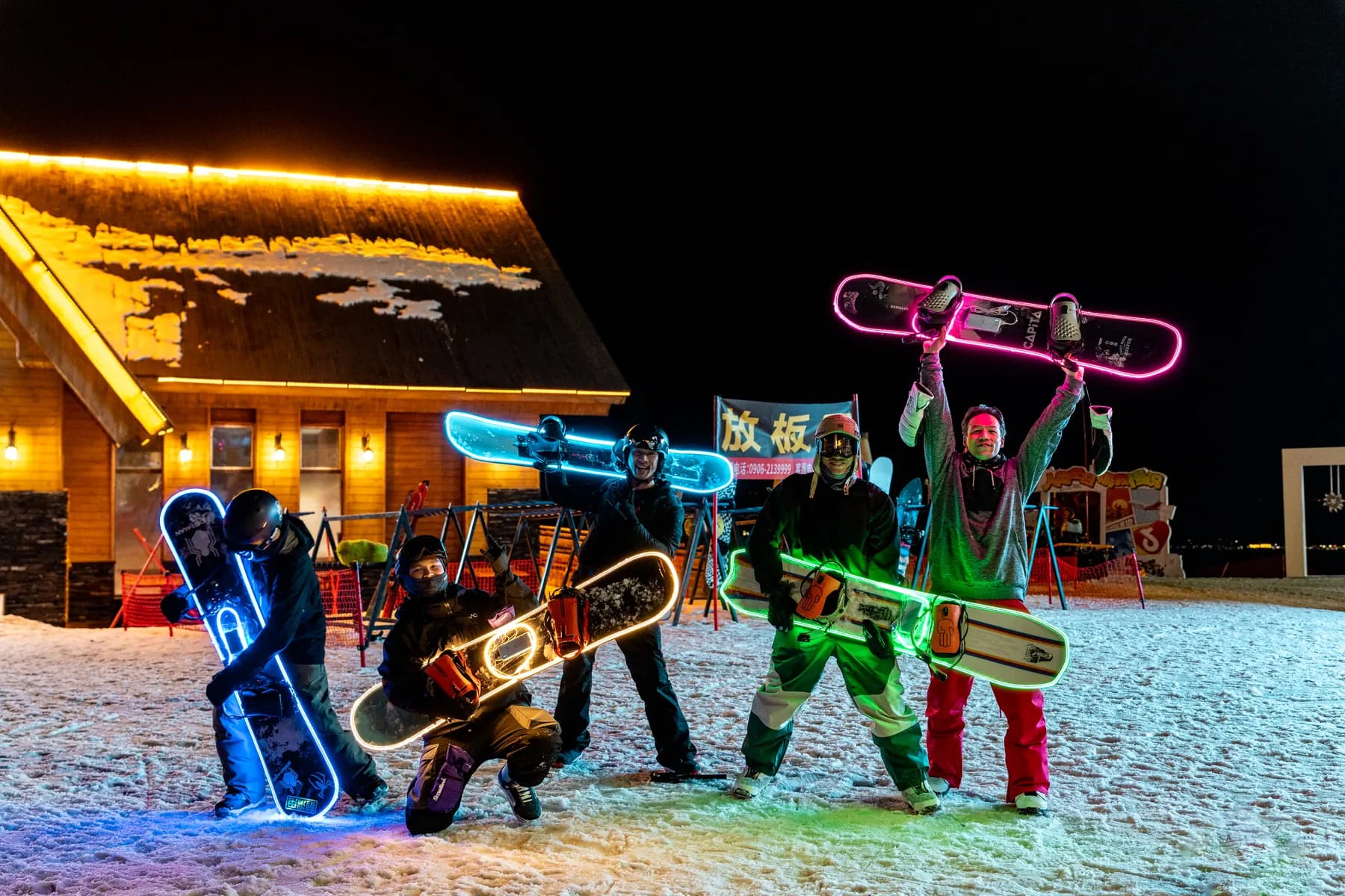 Five people hold illuminated snowboards outside a timber building at night.