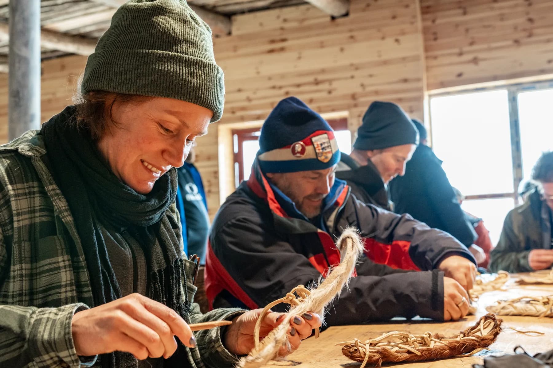 A woman works on a small fur-covered ski model at a wooden table, with other people beside her.