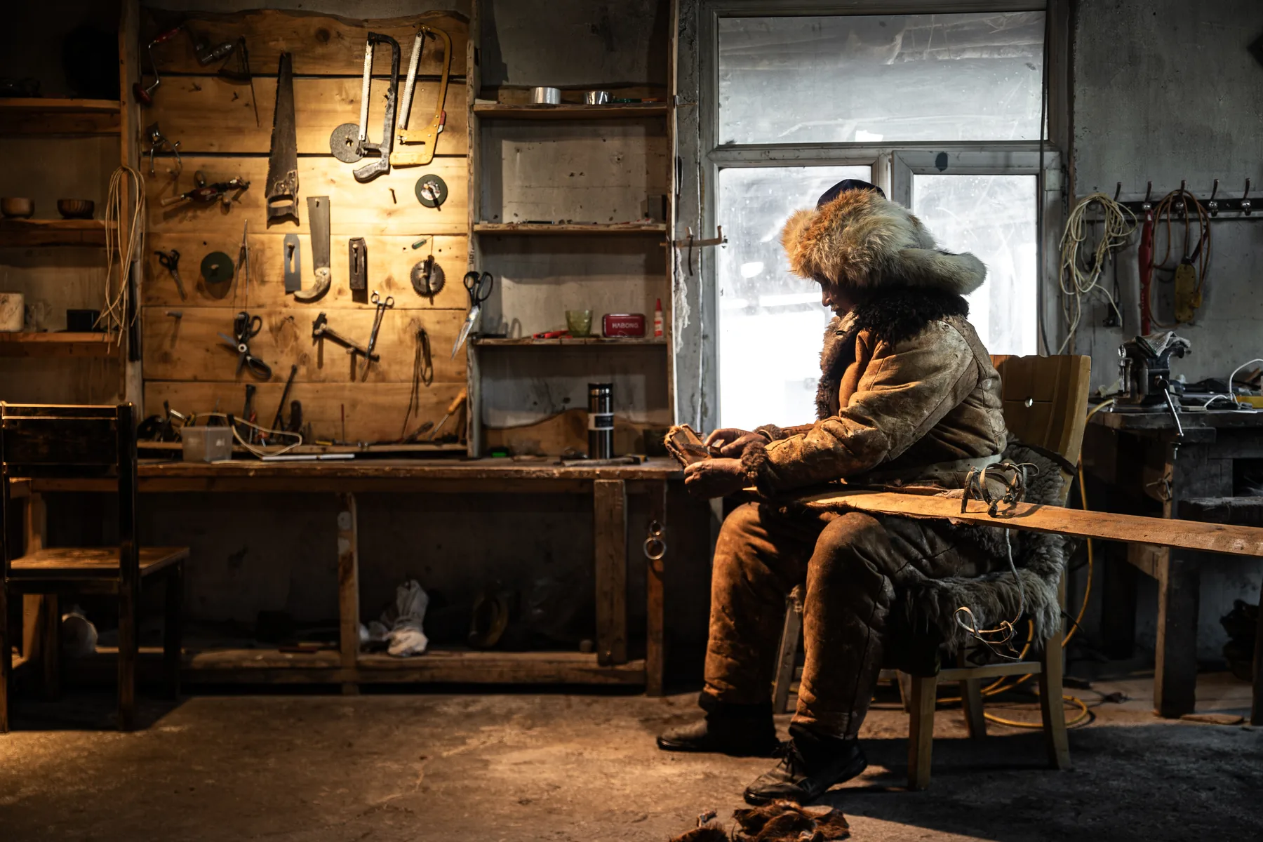 A person in a fur-trimmed coat sits beside a wall of tools in a timber workshop.
