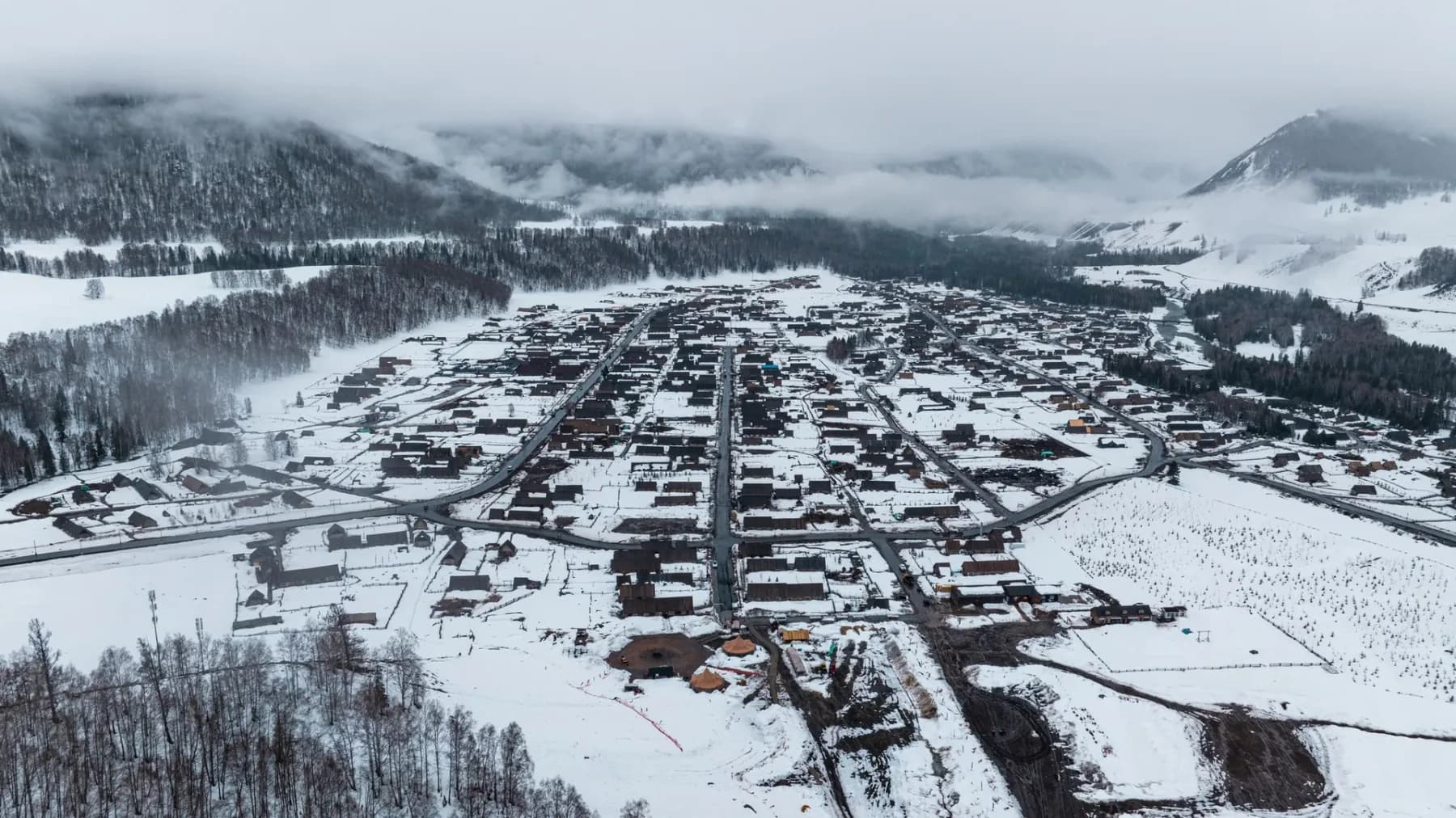 The village of Hemu from the air in deep snow — dark timber cabins and fenced yards spread along a valley floor, with mist hanging in the forest above.