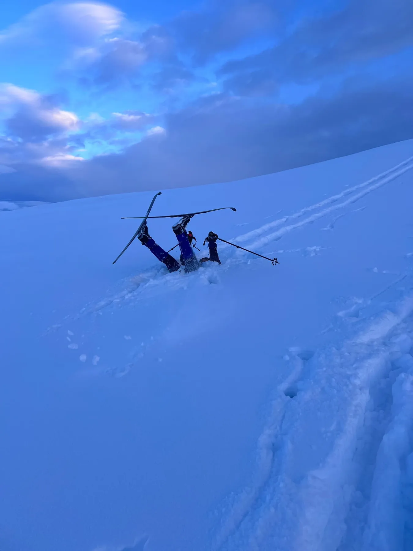 A skier upside down in deep snow with both skis in the air, another skier standing beside them.