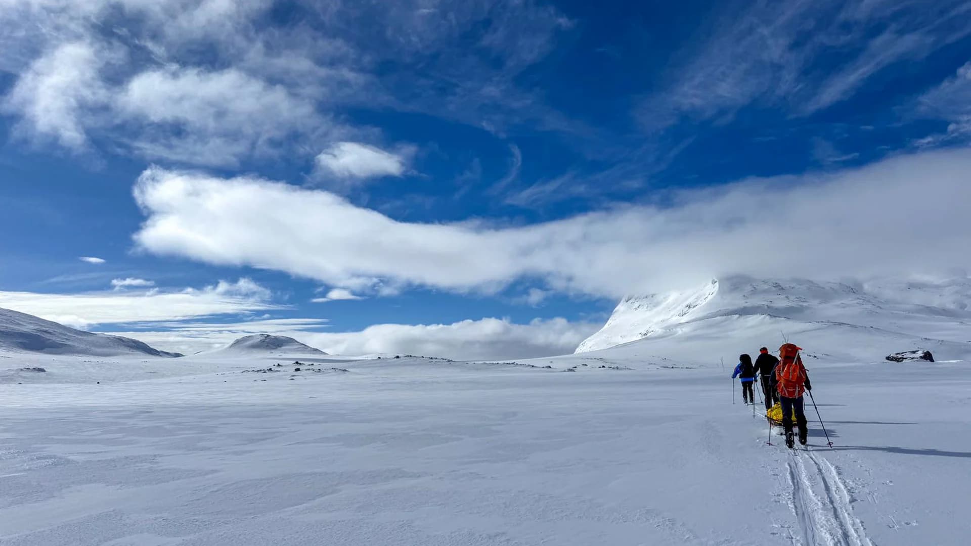 Three skiers hauling a pulk across an open snow plateau under a wide sky, their tracks running back towards the camera.