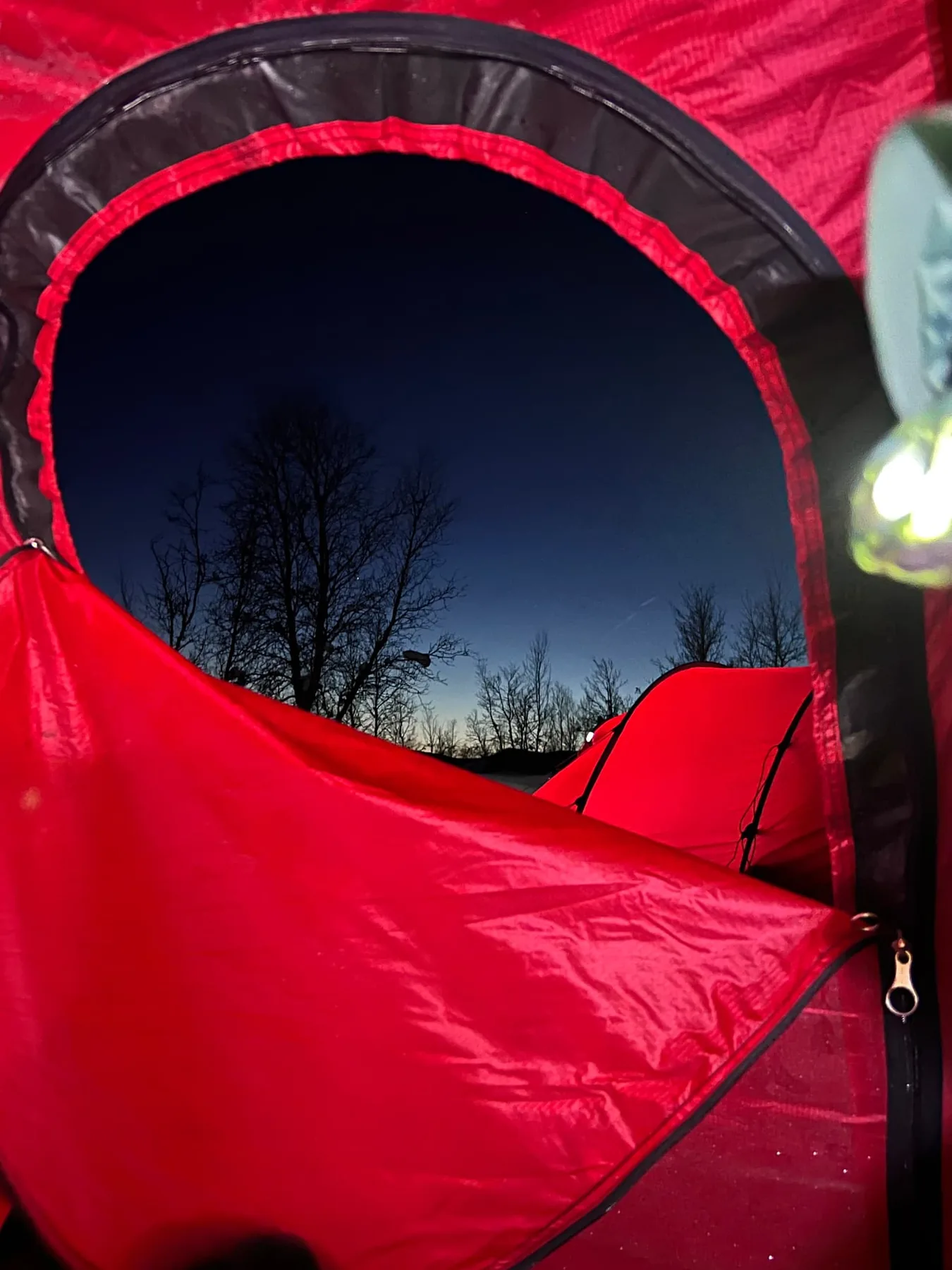 The open round door of a red tent frames bare trees and a deep blue evening sky, with a second red tent pitched outside.