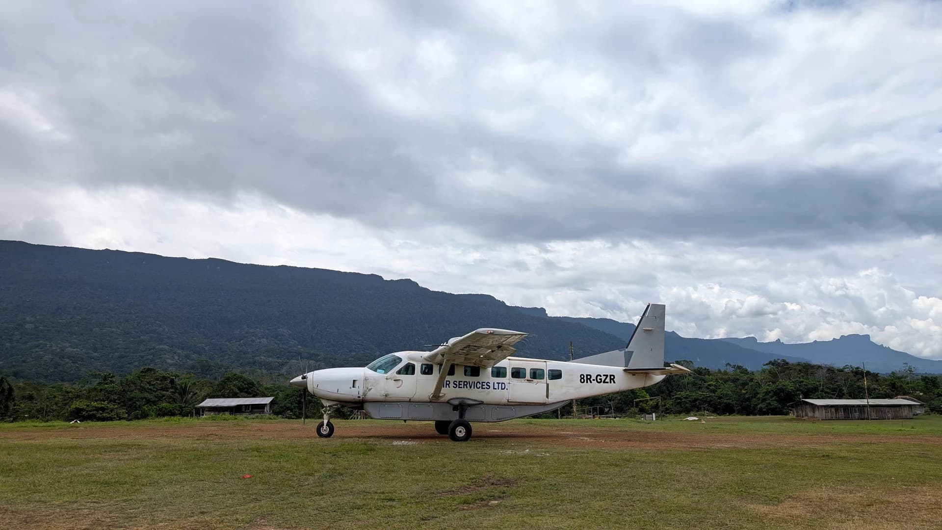 A single-engined Cessna Caravan parked on a grass airstrip, with the forested tepui escarpment running along the horizon behind it.