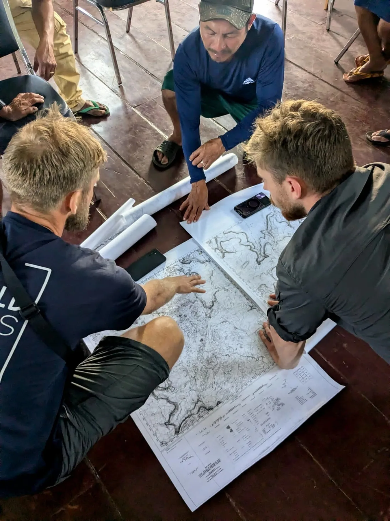 Three people crouched over large printed topographic map sheets spread on a wooden floor, rolled maps beside them.