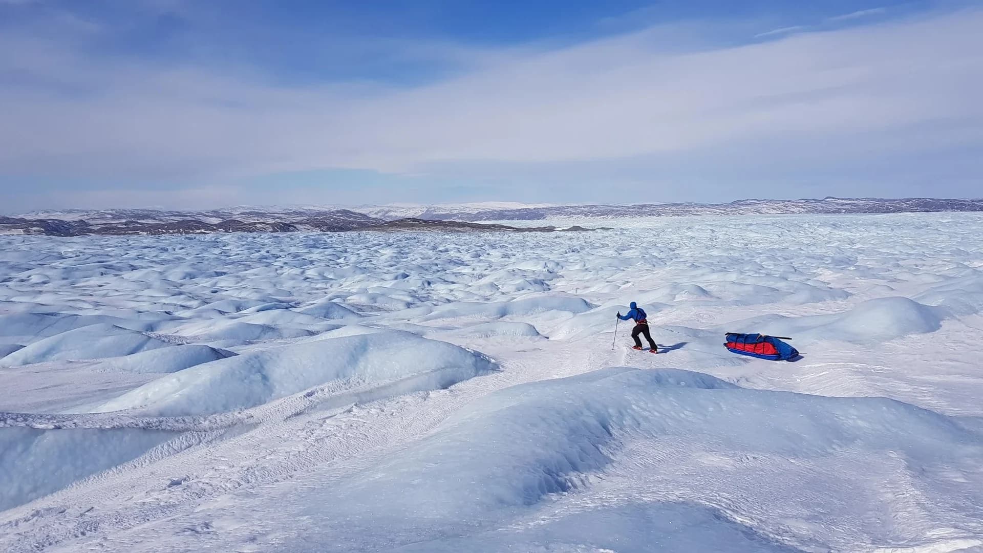 A traveller pulling a pulk across a wide, wind-shaped snowfield.