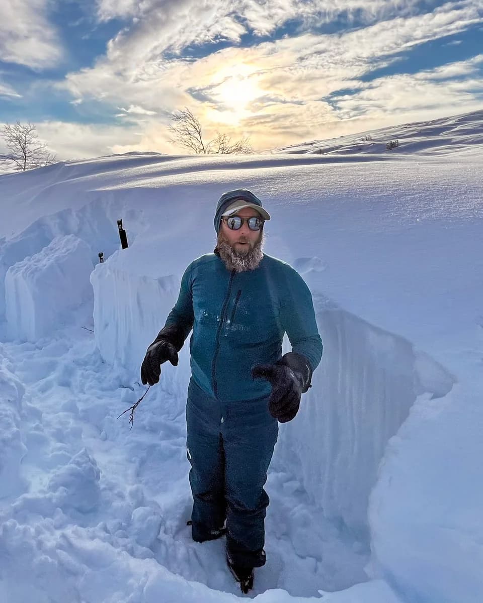 Jamie standing in a trench cut into deep snow, frost in his beard, with a low sun behind him.