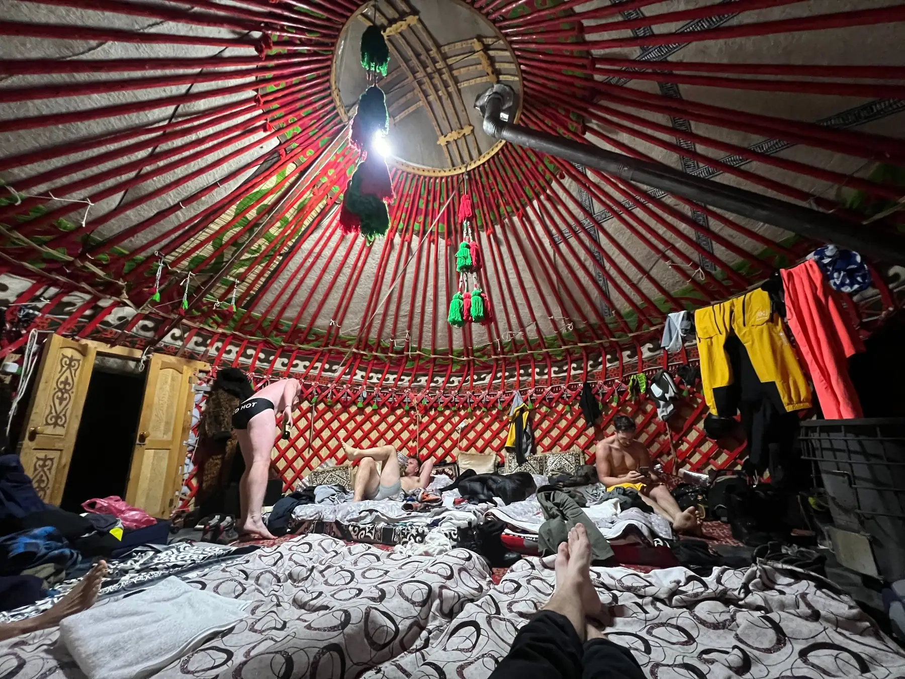 Inside a yurt with a red painted lattice wall and roof poles, several people lying on bedding across the floor, outer kit hung up to dry along the wall.