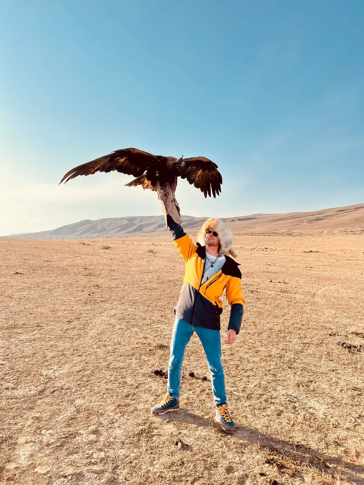 A traveller in a yellow jacket and fur hat holding an eagle above his head on a gloved hand, its wings fully spread, flat dry plain in every direction.