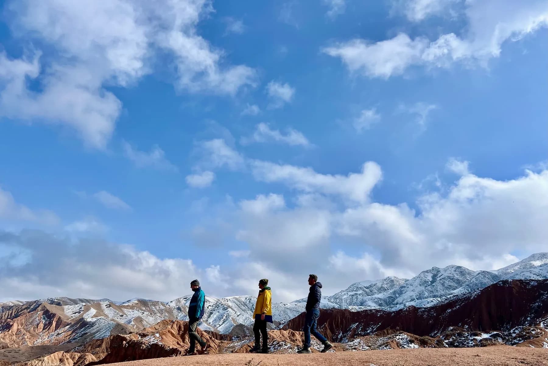 Three people in jackets and hats walking along a bare red earth ridge, snow-dusted mountains and a wide cloudy sky behind.