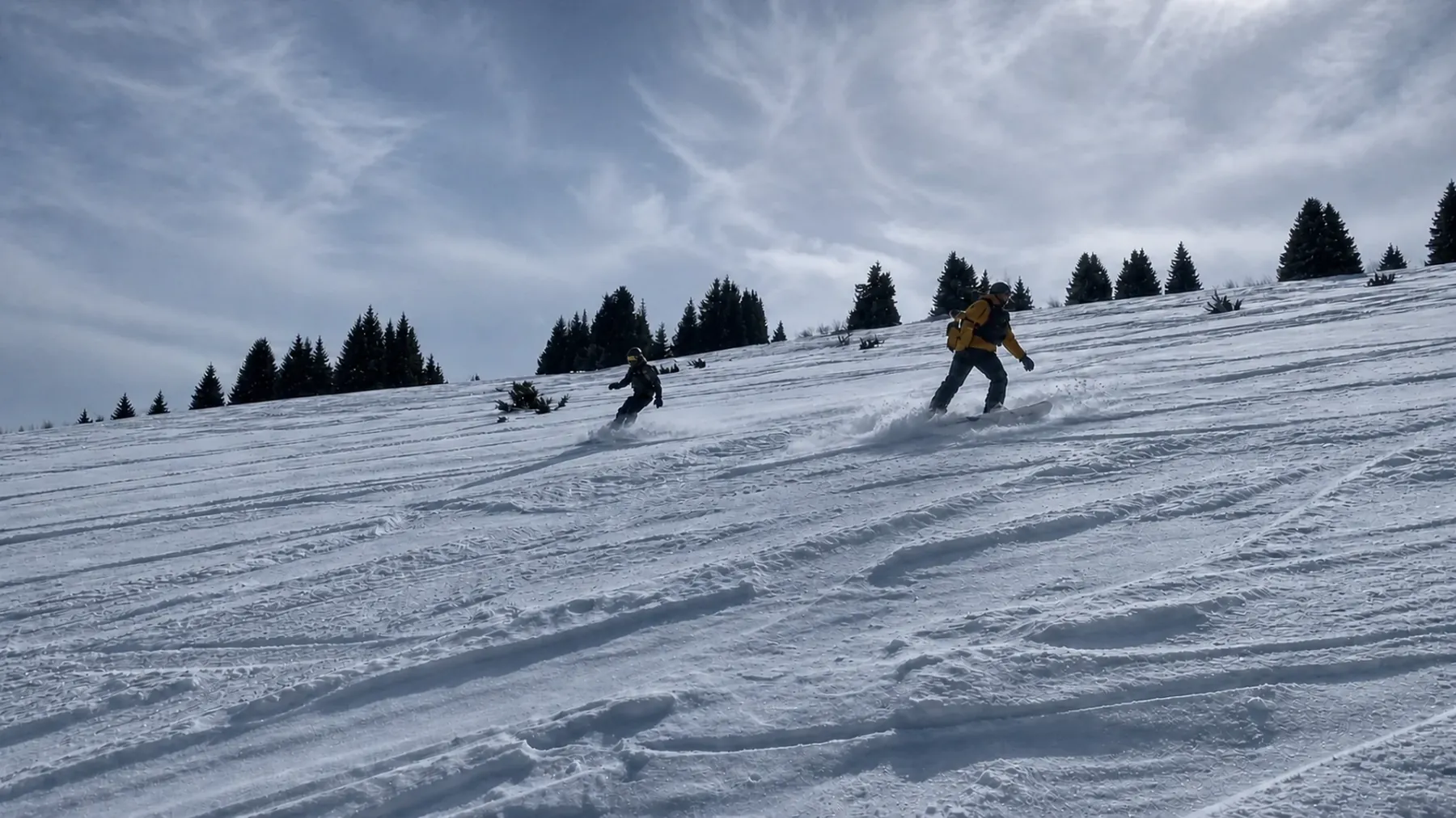 Two riders on a tracked snow pitch below a treeline, fresh lines cut across the slope.