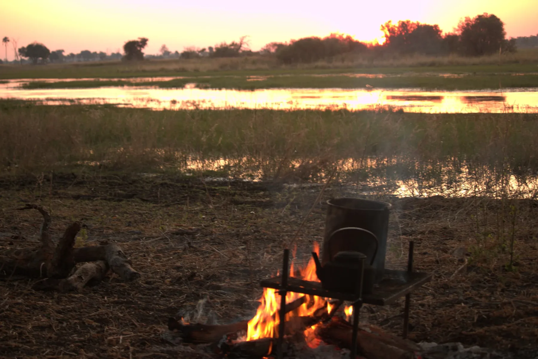 A small campfire burns under a metal pot on a stand, beside flooded grassland reflecting a low orange sun.
