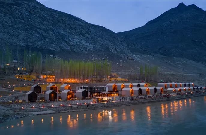 Lit cabins beside a river at dusk, beneath a steep bare mountain.