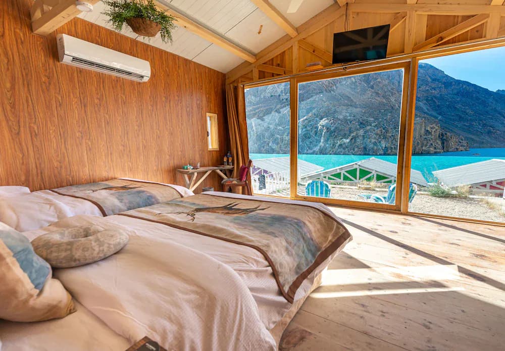 A wood-lined bedroom with broad windows looking onto turquoise water and mountains.