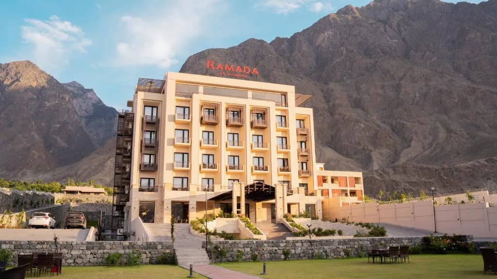 A cream hotel building and lawns at the foot of a high rocky ridge.