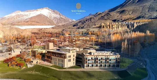 A low stone hotel, lawns and autumn trees in a broad valley below snow-covered mountains.
