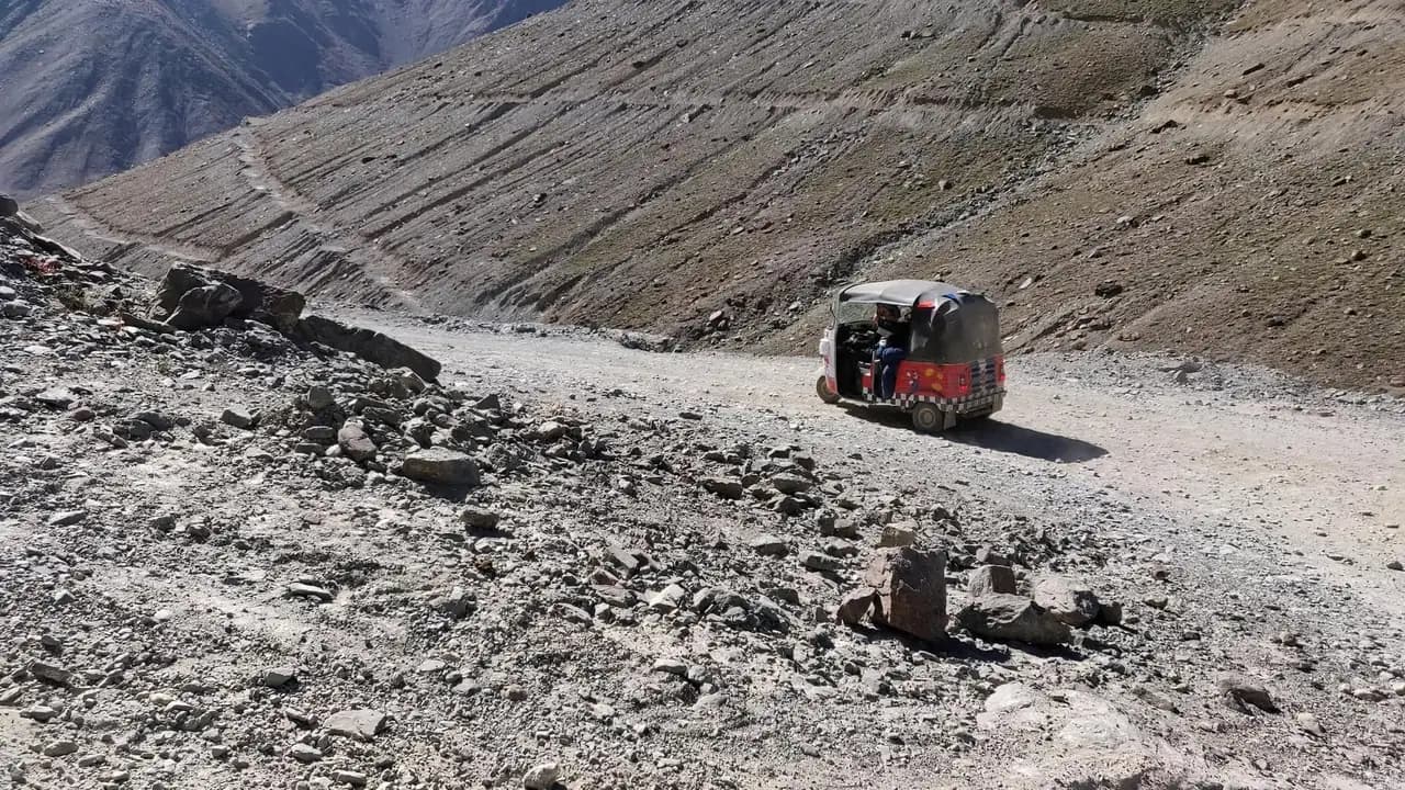 A three-wheeled vehicle on a rough gravel road below a huge grey scree slope, with fallen rock scattered across the track in the foreground and switchback lines cut across the hillside above.