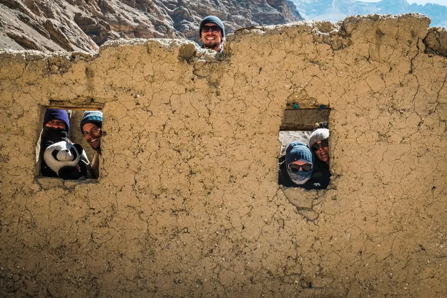 Five people in wool hats and sunglasses look out through two window openings and over the top of a cracked mud-brick wall. One of them holds a large soft toy panda.