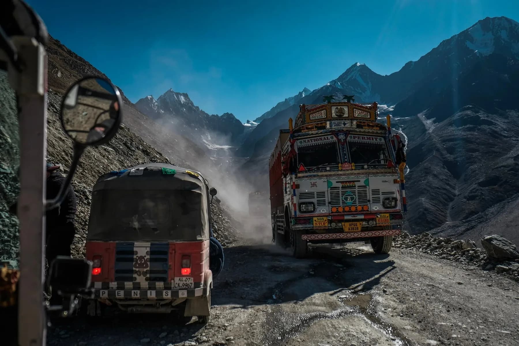 Seen past a wing mirror, a three-wheeled vehicle and a heavily decorated cargo truck pass each other on a dusty mountain road below snow-covered peaks.