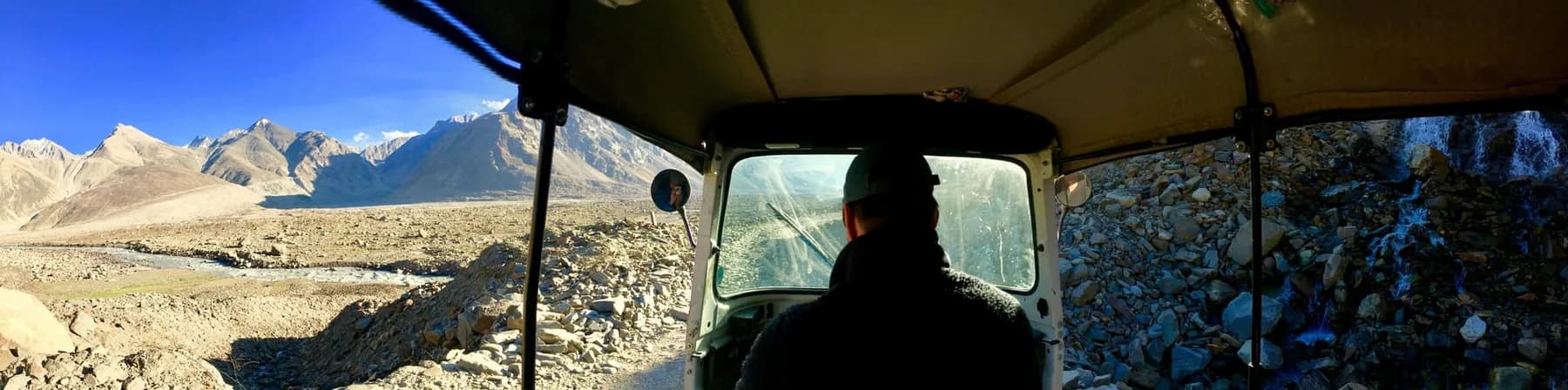 A wide view from inside a three-wheeler, with the driver's silhouette at the wheel, a boulder-strewn riverbed and mountains to the left and rock close on the right.