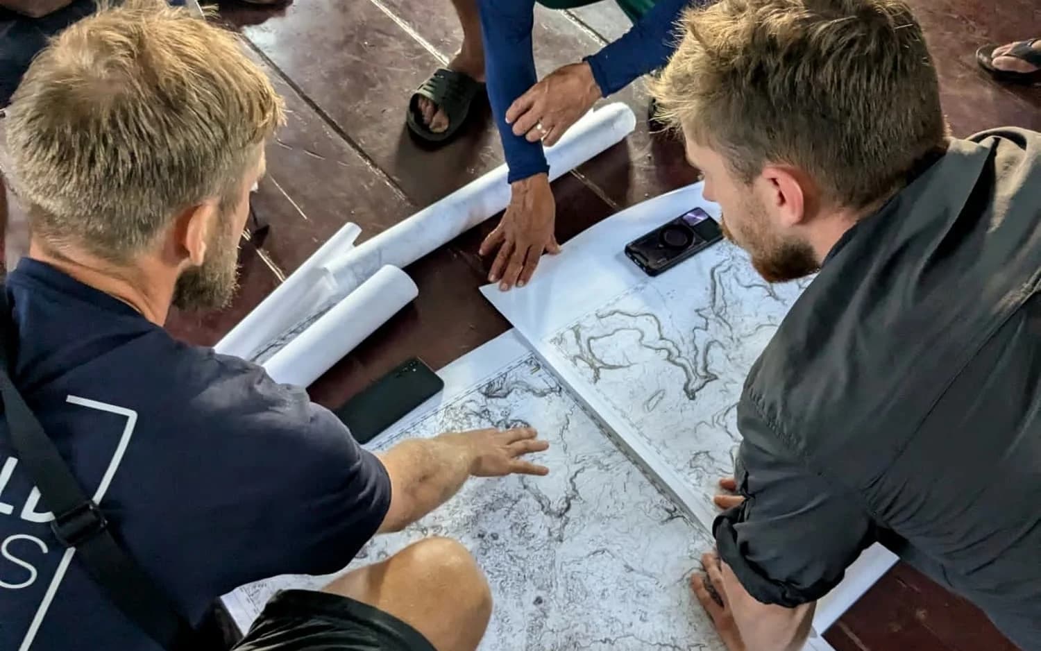 A local guide's hand resting on rolled charts while two men lean over a large printed topographic map on a wooden floor.