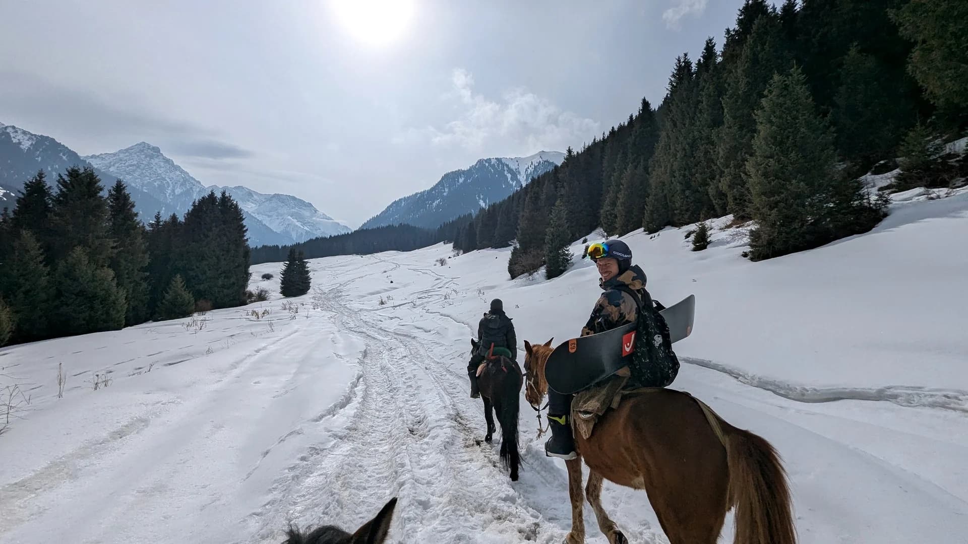 Riders on horseback crossing a snowfield towards forested mountains, one turning in the saddle with a snowboard strapped across his back.
