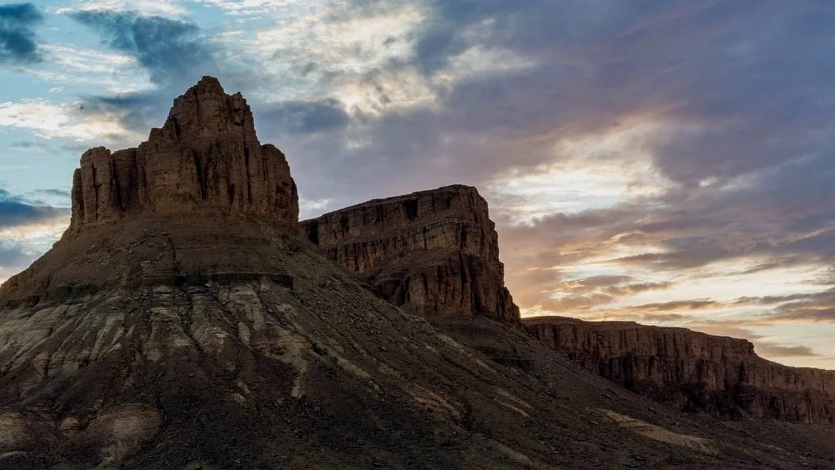A layered sandstone butte with a broken pinnacle on top, catching low light above dark scree slopes, with a long cliff line running away to the right under a streaked evening sky.