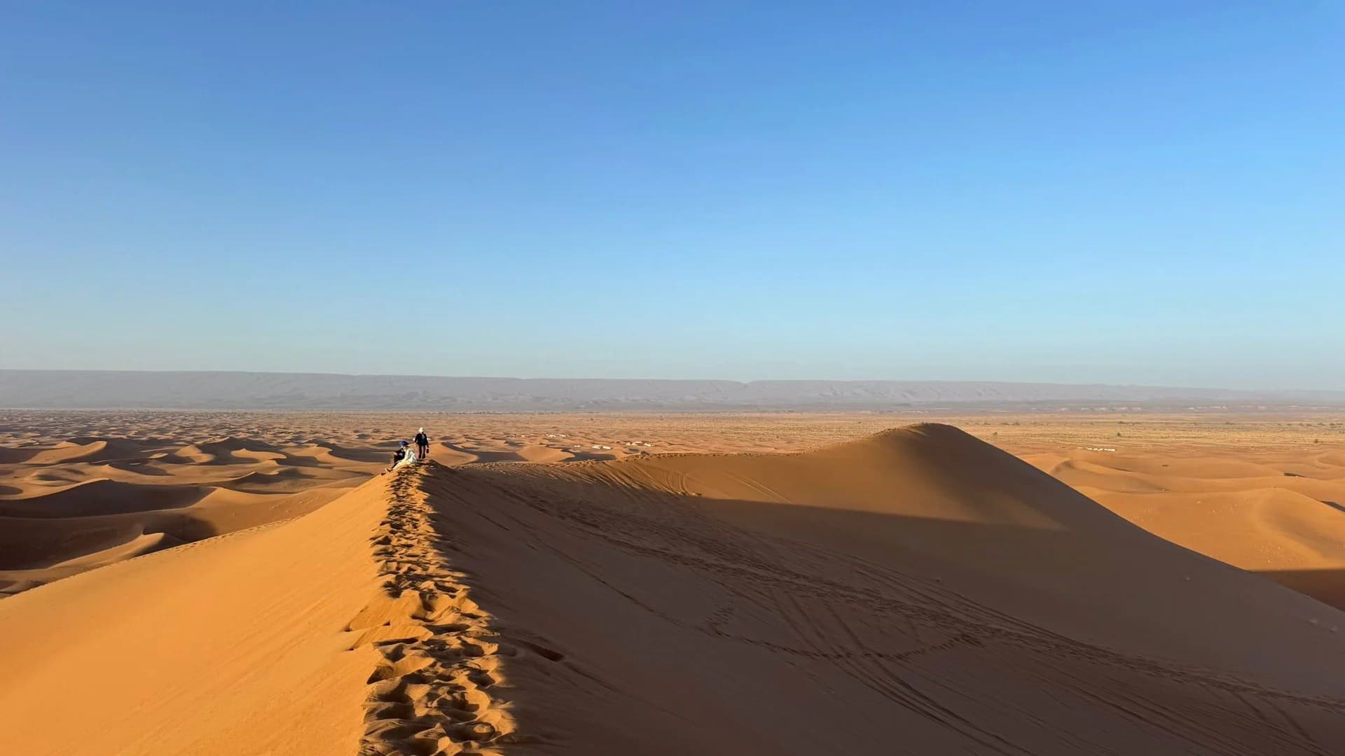 Two walkers on the crest of a dune at low sun, footprints running back down the ridge towards the camera.