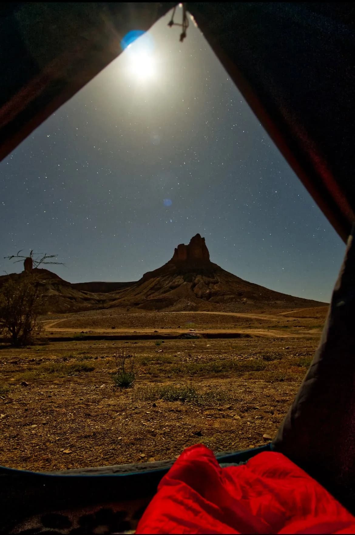 The view out of a tent door at night, a bright moon and stars above a steep-sided butte, a red sleeping bag in the foreground.