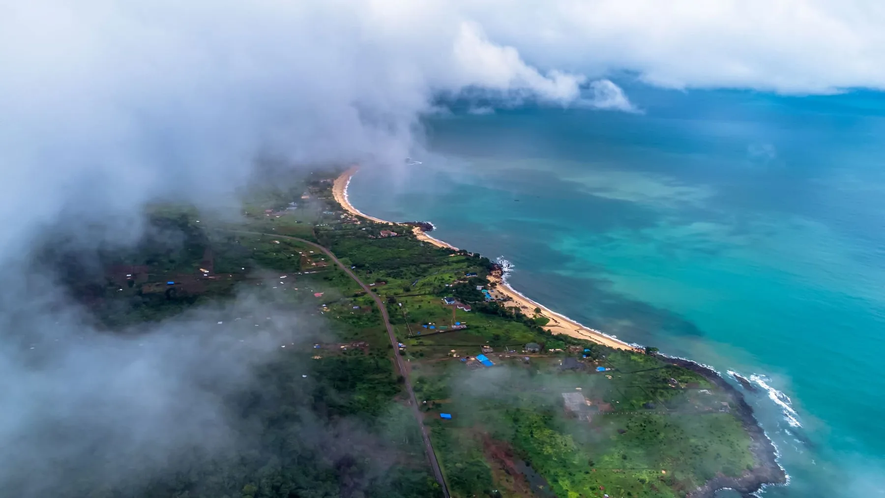 The Freetown peninsula from the air through broken cloud: turquoise shallows, a pale beach and a road running the length of a green headland.
