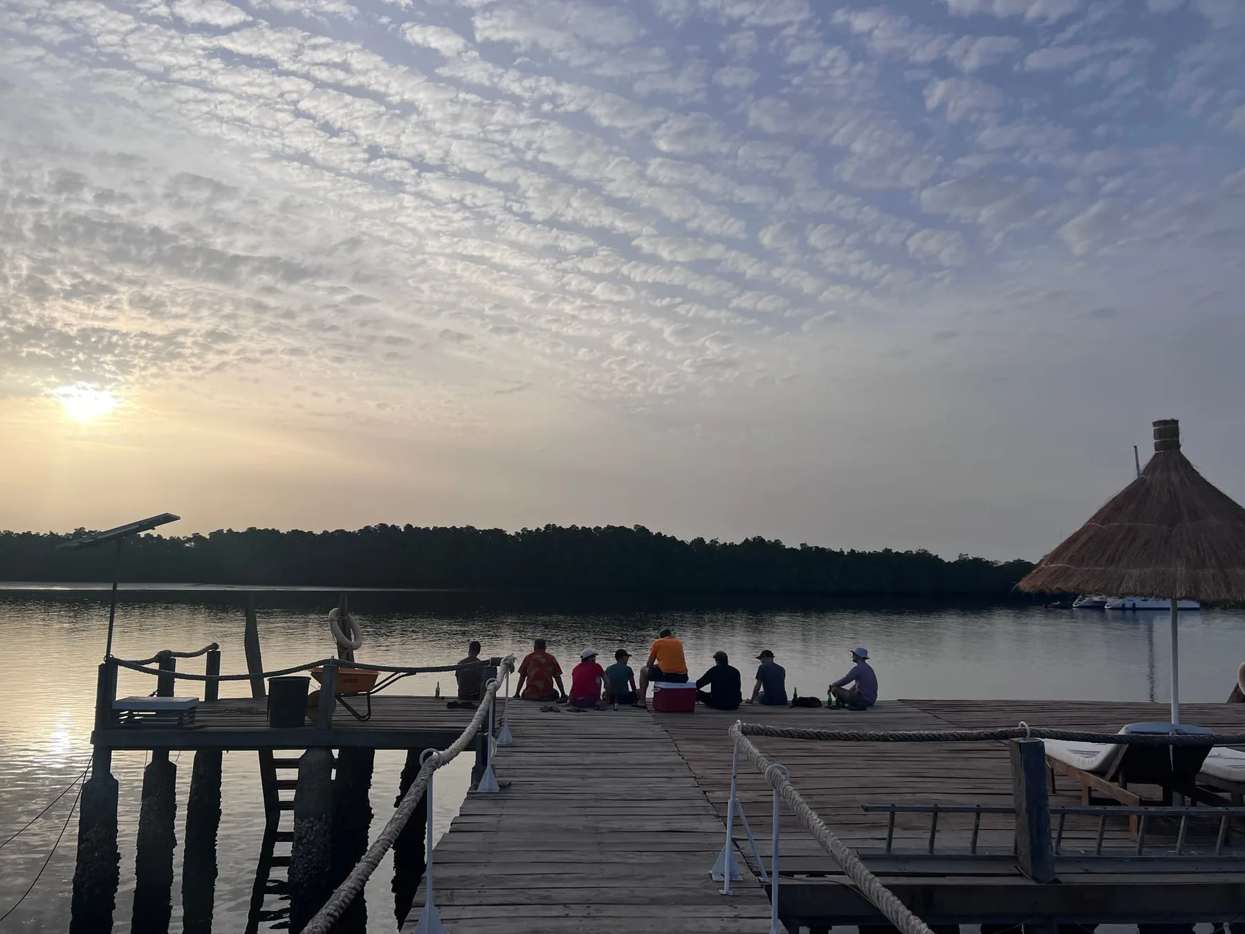 Eight or nine people sit in a row at the end of a wooden jetty at sunset, a red cool box beside them.