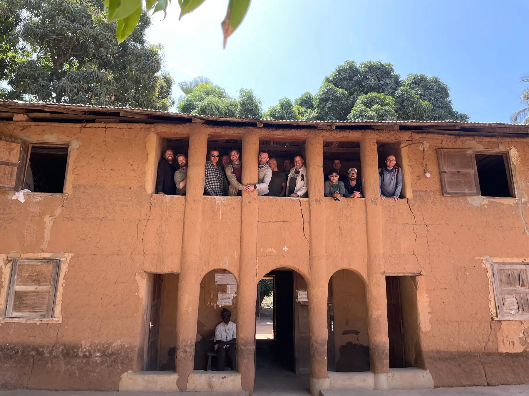About a dozen people lean out from the upper-floor openings of a two-storey earthen building. A man sits in an archway below.