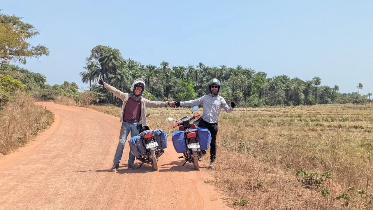 Two motorcyclists standing beside loaded bikes on a red dirt road