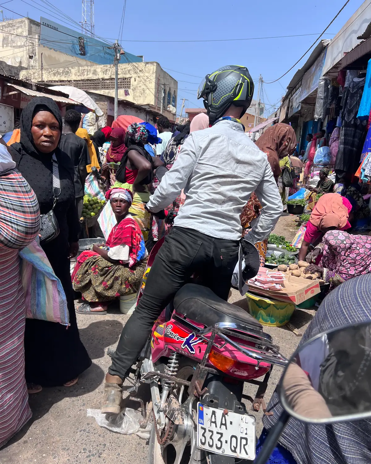 A rider in a helmet sits on a pink motorbike wedged in a crowded market street, with stallholders, shoppers and produce laid out on the ground either side.