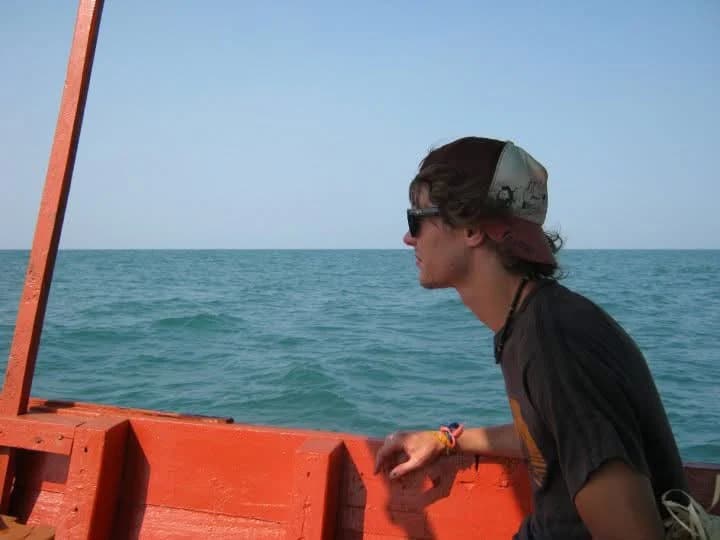 A young man in sunglasses and a backwards cap leans on the red-painted side of a wooden boat, looking out across open blue-green sea to a flat horizon.