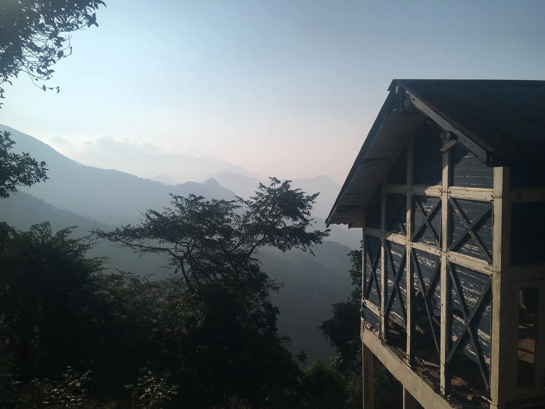 The gable end of a raised wooden building with a steep dark roof, seen through trees on a hillside with layers of misty forested mountains behind it.