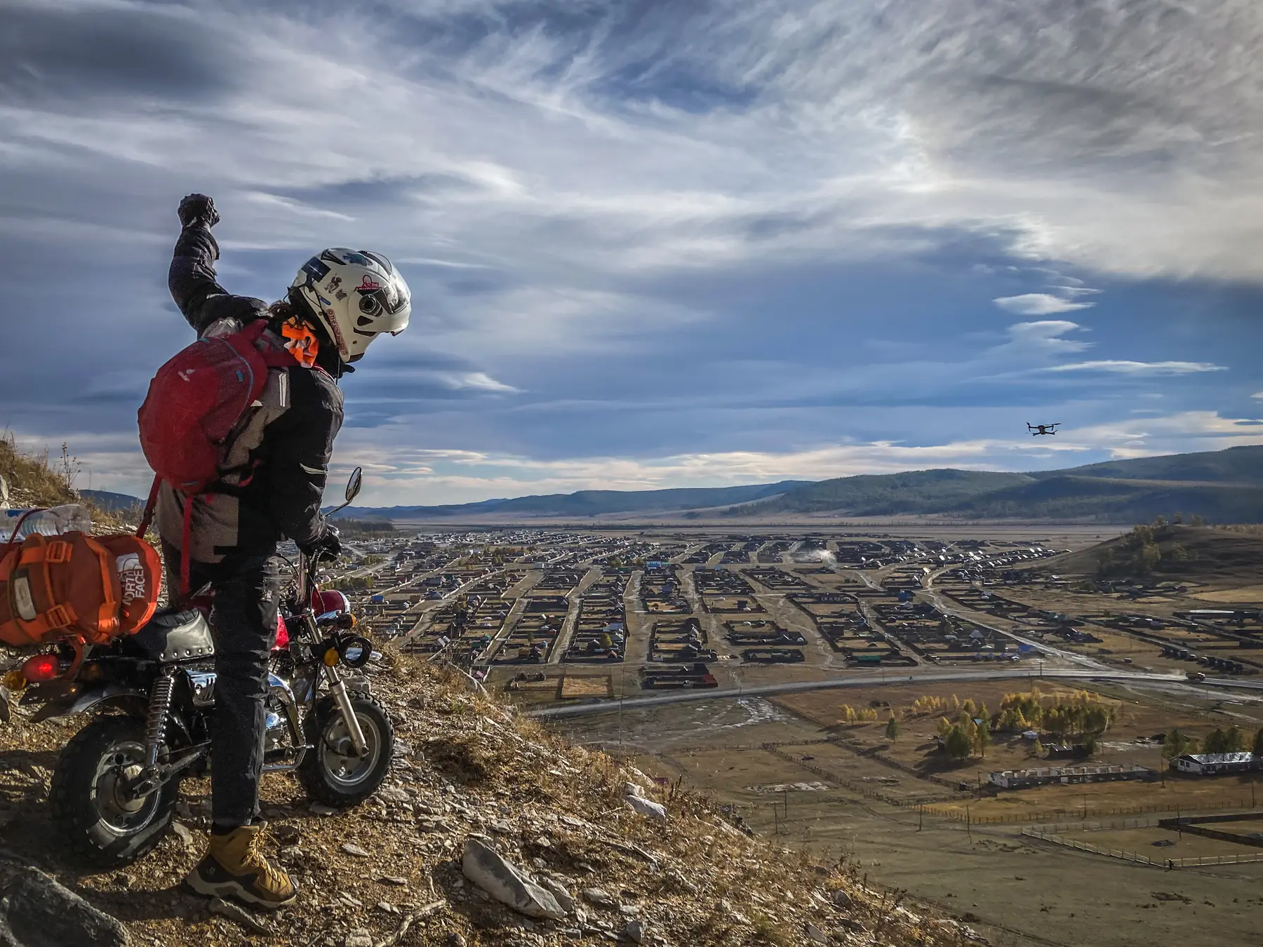 A rider standing over a small off-road motorbike with one fist raised, looking down over a grid of walled housing plots filling a wide valley, hills beyond and a small drone hovering in the middle distance.