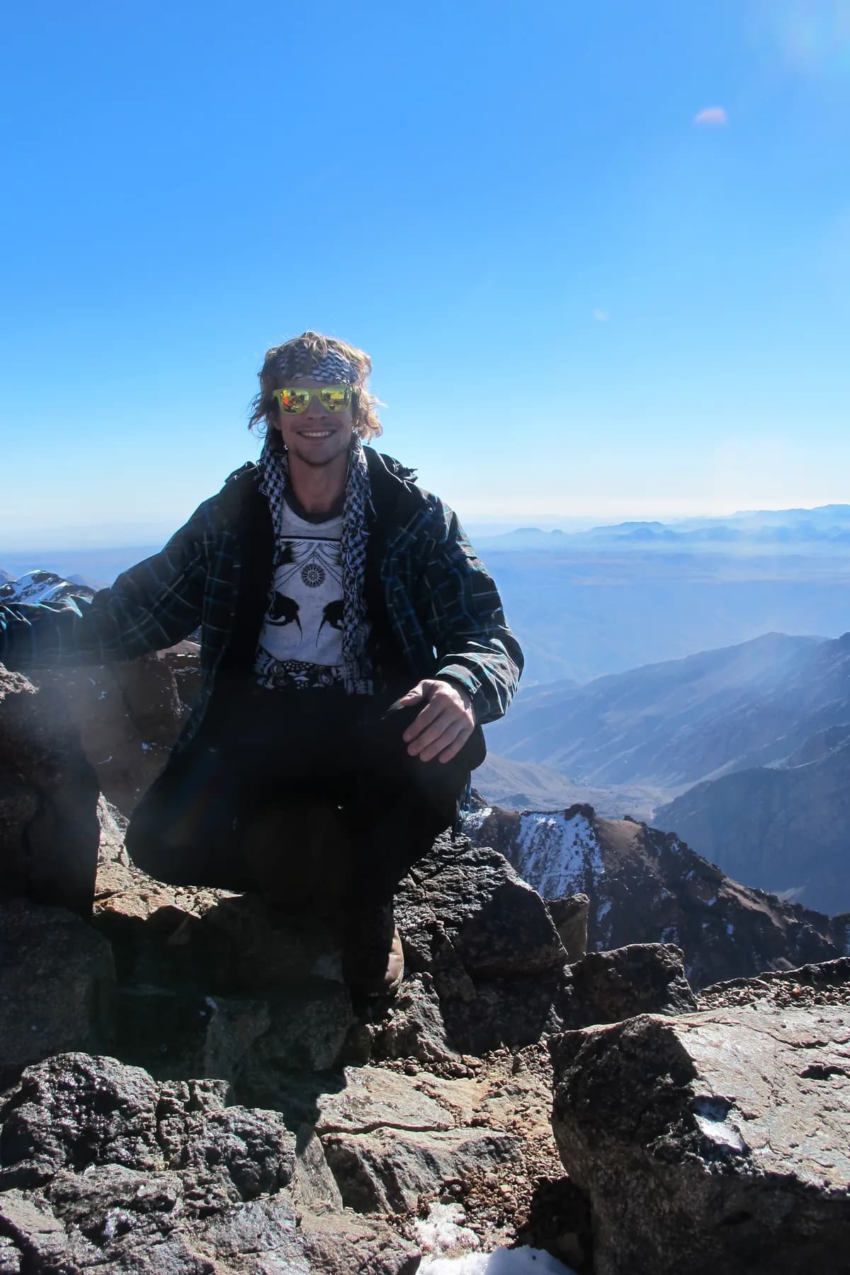 A man in a chequered scarf and sunglasses sitting on dark rock, patches of snow around him and layered mountains in the distance.