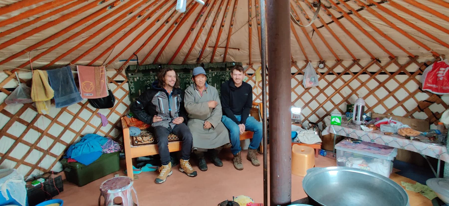 Three men sit posed on a wooden bench inside a fabric-lined yurt, storage boxes, towels and a kettle around them.