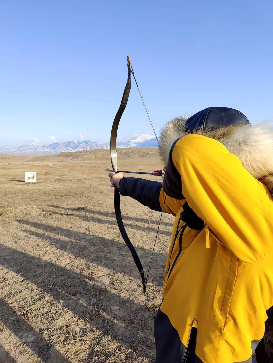 Drawing a Kyrgyz horn bow on the open steppe, a deer target in the distance and snow peaks beyond.