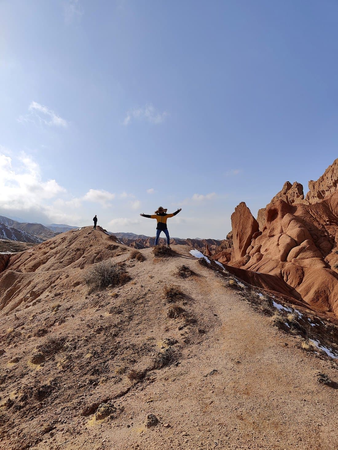Standing on a ridge above the red rock badlands of Fairy Tale Canyon, arms out, a second walker on the skyline behind.